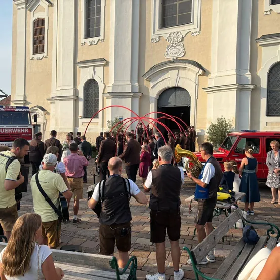 Eine Gruppe von Menschen versammelt sich vor einer Kirche, einige spielen Musikinstrumente, mit einer roten Struktur im Vordergrund und einem roten Fahrzeug in der Nähe.