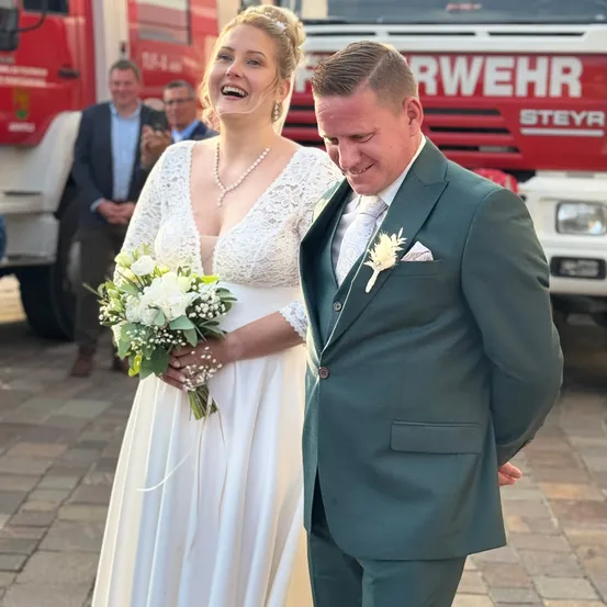 Eine Braut in einem weißen Kleid und ein Bräutigam in einem grünen Anzug lächeln und gehen vor einem Feuerwehrauto.