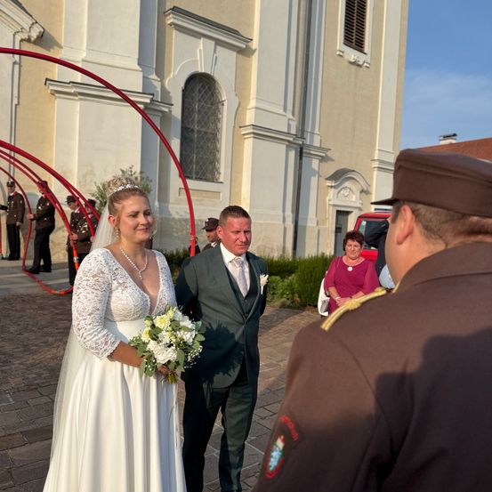Ein frisch verheiratetes Paar verlässt eine Kirche mit einem Offizier; die Braut hält Blumen, und der Bräutigam trägt eine Krawatte. Hinter ihnen stehen Soldaten Wache.