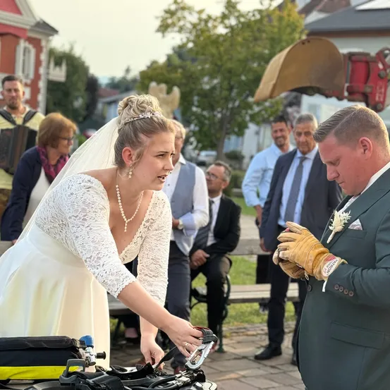 Eine Braut in einem weißen Spitzenkleid und Schleier steht mit dem Rücken zur Kamera und hält ein Werkzeug. Ein Bräutigam in einem grünen Anzug und gelben Handschuhen schaut sie an. Andere Leute sind im Hintergrund, einschließlich eines Mannes mit einem Rucksack.