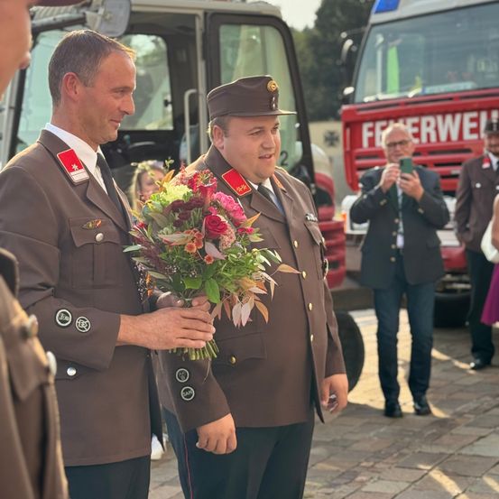 Zwei uniformierte Männer halten einen Blumenstrauß draußen. Sie stehen vor einem Feuerwehrauto mit dem Wort Feuerwehr. Ein Mann hinter ihnen macht ein Foto mit dem Handy.