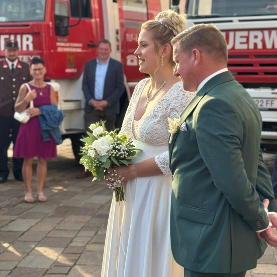 Ein frisch verheiratetes Paar steht nah beieinander und lächelt, mit einem Brautstrauß in der Hand der Braut. Dahinter ist ein roter Feuerwehrwagen geparkt, und mehrere Personen stehen herum.