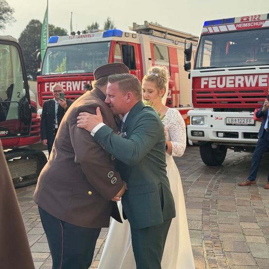 Ein Mann in Militäruniform umarmt einen Mann in einem grünen Anzug, während eine Frau in einem weißen Kleid zusieht. Hinter ihnen stehen Feuerwehrfahrzeuge.