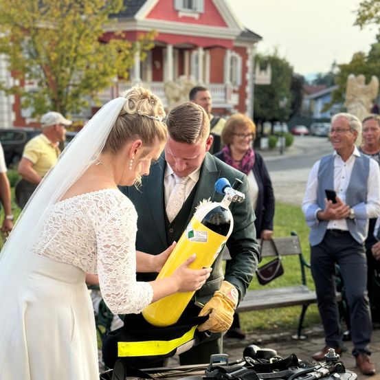 Ein Hochzeitspaar hält an ihrem Hochzeitstag eine Tauchflasche in der Hand, während sie Schutzausrüstung tragen. Hinter ihnen beobachtet eine kleine Menge. Im Hintergrund ist ein Haus und einige geparkte Autos zu sehen.