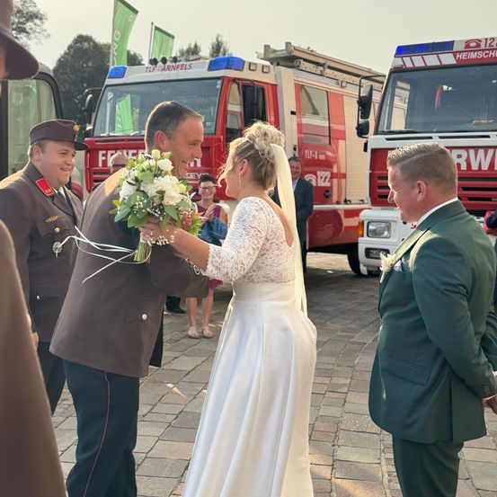 Eine Hochzeitszeremonie mit der Braut und dem Bräutigam vor einer Gruppe von Feuerwehrleuten mit Feuerwehrautos im Hintergrund.