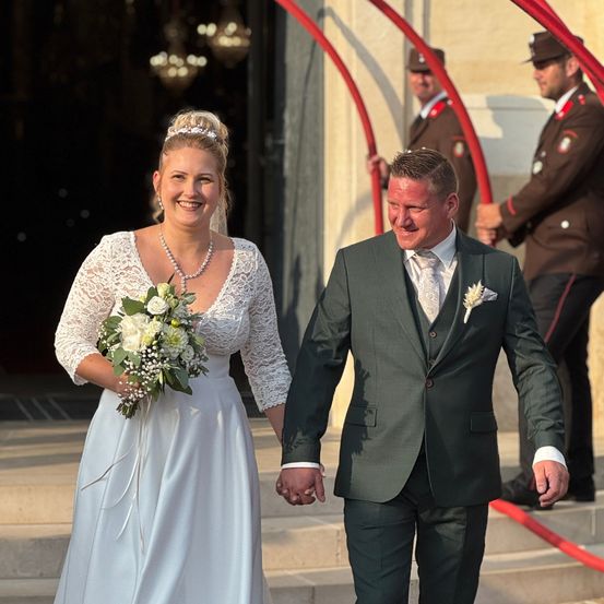 Eine Braut und ein Bräutigam gehen lächelnd die Stufen hinunter. Die Braut hält einen Blumenstrauß. Hinter ihnen stehen zwei Männer in Militäruniformen. Die Szene spielt sich im Freien ab.