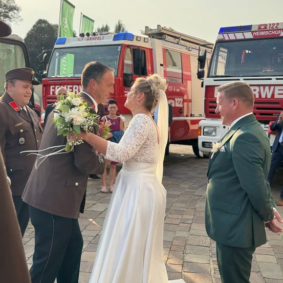 Eine Hochzeitszeremonie mit einer Braut in einem weißen Kleid und einem Bräutigam im Anzug, umgeben von Feuerwehrleuten in Uniform. Sie stehen vor Feuerwehrwagen.