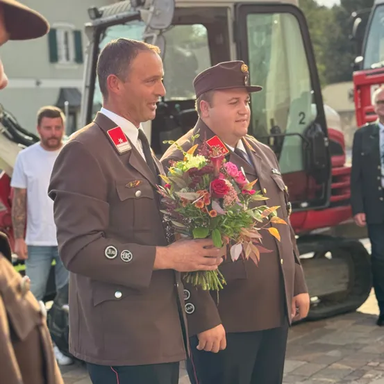 Zwei Männer in braunen Uniformen halten einen Blumenstrauß und lächeln. Hinter ihnen steht ein großes rotes Baufahrzeug. Im Hintergrund steht ein Mann in einem weißen T-Shirt und Jeans neben dem Fahrzeug.