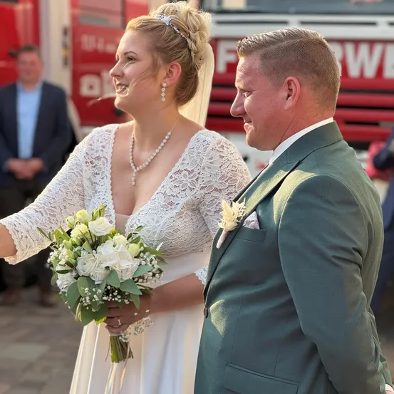 Ein frisch verheiratetes Paar steht draußen, die Braut in einem weißen Kleid, eine Blumenstrauß haltend, und der Bräutigam in einem grünen Anzug. Sie lächeln sich an. Hinter ihnen ist ein Feuerwehrauto zu sehen.