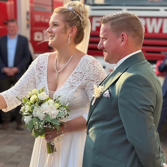 Ein frisch verheiratetes Paar steht draußen, die Braut in einem weißen Kleid, eine Blumenstrauß haltend, und der Bräutigam in einem grünen Anzug. Sie lächeln sich an. Hinter ihnen ist ein Feuerwehrauto zu sehen.