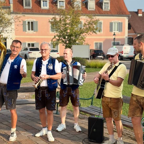 Vier Männer in traditioneller bayerischer Kleidung spielen auf einem Stadtplatz, einer mit Trompete, ein anderer mit Akkordeon und die anderen mit Gitarren.