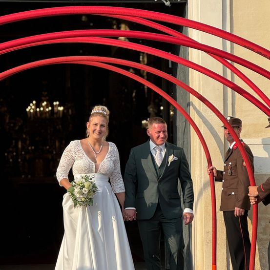 Eine Braut in einem weißen Hochzeitskleid und ein Bräutigam in einem schwarzen Anzug gehen Hand in Hand. Sie sind von roten Bögen umgeben.