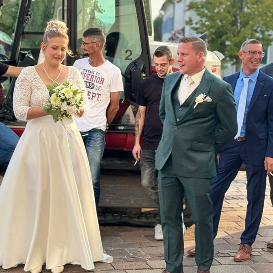 Eine Braut in einem weißen Kleid hält einen Blumenstrauß, während sie neben einem Bräutigam in einem grünen Anzug steht. Dahinter schaut ein Mann in einem weißen T-Shirt und Jeans zu, und ein anderer Mann in einem blauen Anzug lächelt.