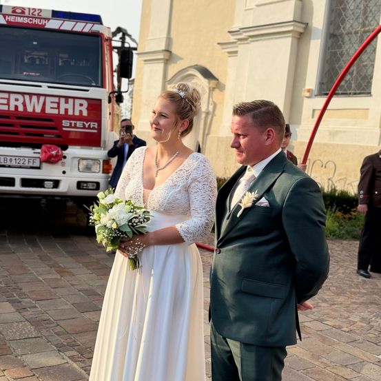 Ein frisch verheiratetes Paar steht draußen, die Braut in einem weißen Kleid und der Bräutigam in einem grünen Anzug. Im Hintergrund steht ein Feuerwehrwagen, und ein Mann macht ein Foto.