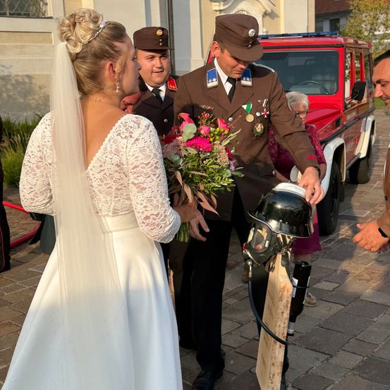 Eine Braut in einem weißen Kleid und Schleier hält einen Blumenstrauß, während ein Offizier ihr einen Helm überreicht. Hinter ihnen steht ein rotes Fahrzeug und ein Mann in einem Anzug.