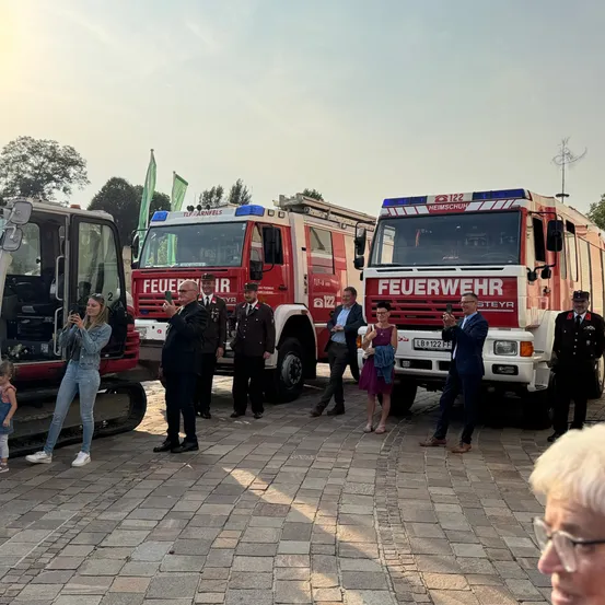 Eine Gruppe von Menschen steht vor Feuerwehrfahrzeugen, einer hält ein Telefon. Eine Frau macht ein Foto, während andere zuschauen. Ein kleines Kind ist anwesend.