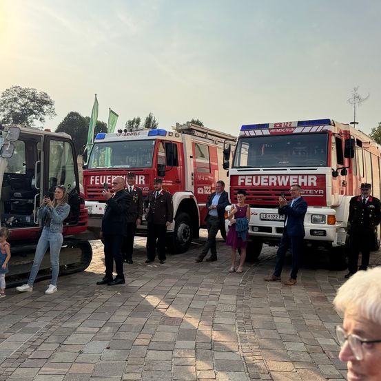 Eine Gruppe von Menschen steht vor Feuerwehrfahrzeugen, einer hält ein Telefon. Eine Frau macht ein Foto, während andere zuschauen. Ein kleines Kind ist anwesend.