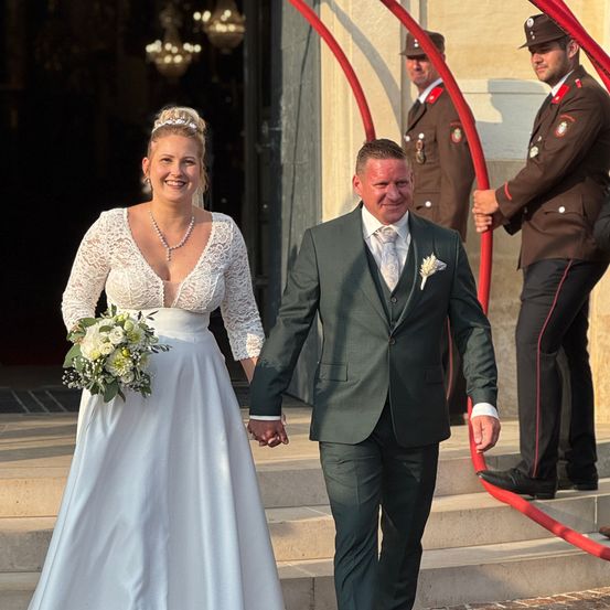 Ein frisch verheiratetes Paar geht eine Treppe hinunter, die Braut in einem weißen Kleid und der Bräutigam in einem grünen Anzug. Sie werden von zwei Männern in Militäruniformen begleitet.