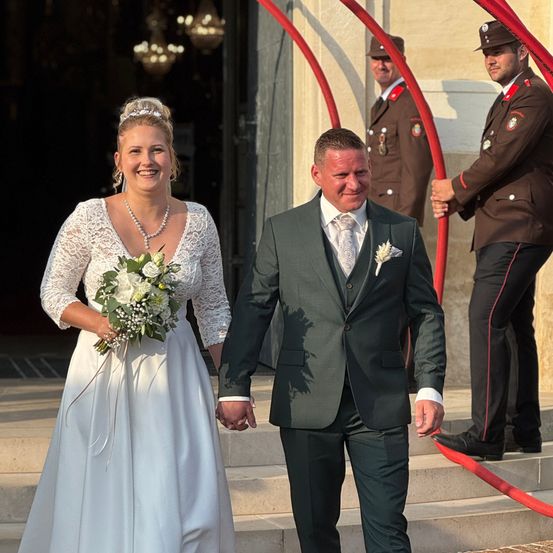Ein frisch verheiratetes Paar, lächelnd, geht eine Treppe hinunter, begleitet von Militärwächtern im Hintergrund.