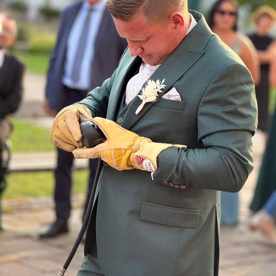 Ein Mann in einem grünen Anzug und Handschuhen hält ein schwarzes Objekt. Er sieht nach unten, möglicherweise bei einer Veranstaltung. Hinter ihm stehen und sprechen Personen.