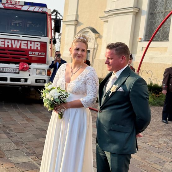 Eine Braut und ein Bräutigam stehen vor einem Feuerwehrauto, die Braut hält einen Blumenstrauß und der Bräutigam trägt einen dunkelgrünen Anzug.