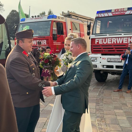 Zwei Männer in Anzügen schütteln sich die Hände vor einem Feuerwehrauto. Eine Frau hält einen Blumenstrauß.