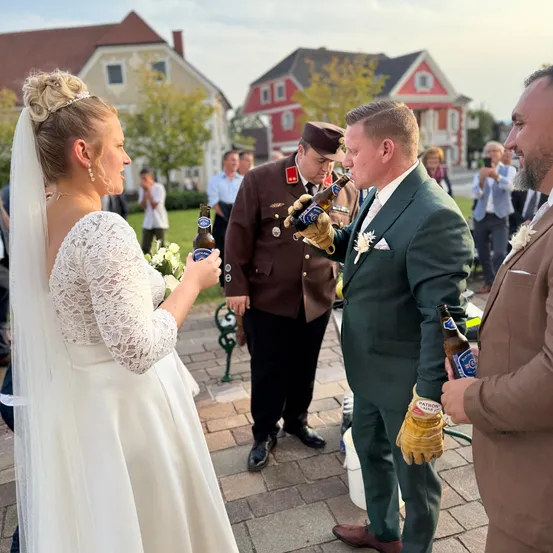 Eine Braut in einem weißen Kleid und ein Bräutigam in einem grünen Anzug öffnen Flaschen mit einem Mann in brauner Uniform mit Handschuhen. Im Hintergrund stehen Menschen in einem kleinen Dorf.