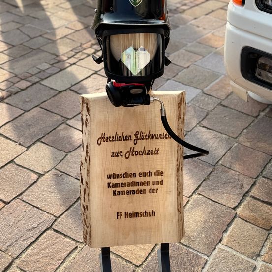 Ein Holzschild mit einem Helm steht auf Kopfsteinpflaster, mit einer Nachricht von FF Heimschuh, die eine glückliche Hochzeit wünscht. Ein Auto ist in der Nähe geparkt.