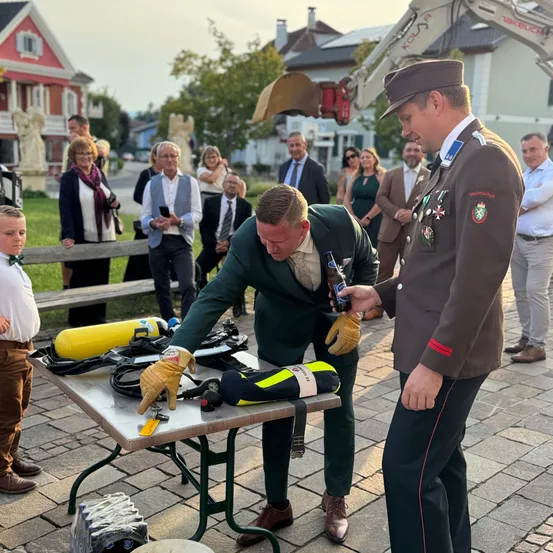 Ein Mann in einem grünen Anzug öffnet eine Bierflasche auf einem Tisch, der von Tauchausrüstung umgeben ist. Ein uniformierter Mann beobachtet, mit Menschen und einem Baufahrzeug im Hintergrund.
