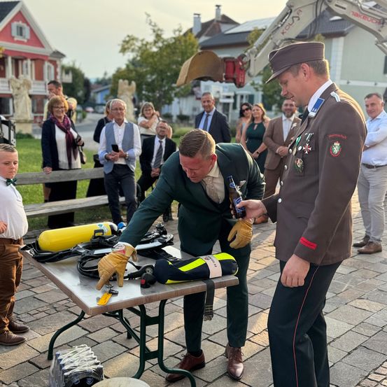 Ein Mann in einem grünen Anzug öffnet eine Bierflasche auf einem Tisch, der von Tauchausrüstung umgeben ist. Ein uniformierter Mann beobachtet, mit Menschen und einem Baufahrzeug im Hintergrund.