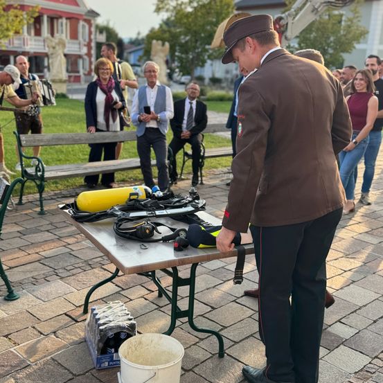 Ein Mann in Uniform steht neben einem Tisch mit Tauchausrüstung. Hinter ihm versammelt sich eine Menschenmenge, einige sitzen auf Bänken. Bäume und Gebäude sind im Hintergrund zu sehen.