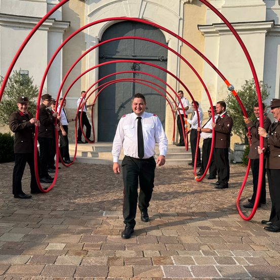 Ein Mann in Uniform geht durch eine rote Bogenformation, die von mehreren Personen vor einem Gebäude gebildet wird.