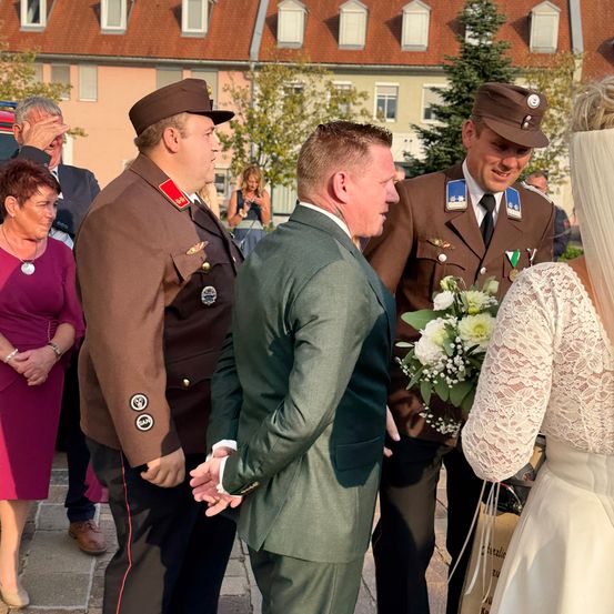 Eine Gruppe von Menschen steht im Freien, darunter zwei Männer in Militäruniformen und ein Mann in einem Anzug, der einen Blumenstrauß hält. Ein Bräutigam und eine Braut stehen eng beieinander, und eine Frau in einem rosa Kleid schaut zu.
