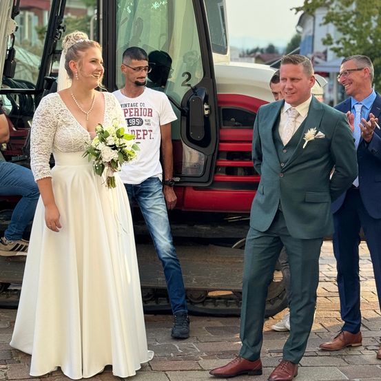 Eine Braut und ein Bräutigam stehen lächelnd vor einem Traktor, während zwei Männer in Anzügen hinter ihnen klatschen. Der Bräutigam trägt einen grünen Anzug und die Braut ein weißes Hochzeitskleid.