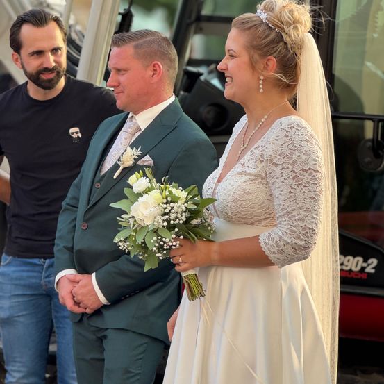 Eine Braut in einem Spitzenhochzeitskleid und ein Bräutigam in einem grünen Anzug stehen lächelnd zusammen, während ein anderer Mann in einem schwarzen T-Shirt zusieht.