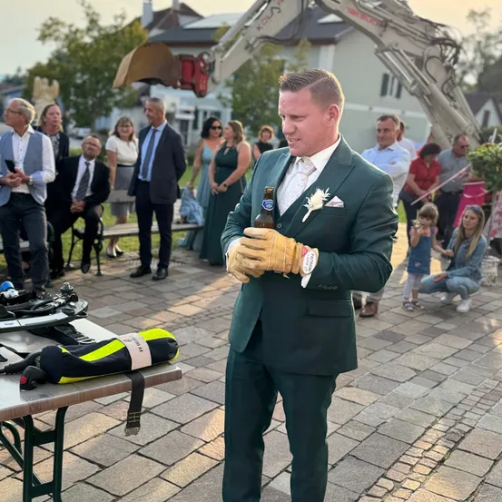 Ein Mann in einem grünen Anzug hält eine Flasche und steht vor einem Tisch mit Schutzausrüstung. Hinter ihm beobachten Menschen, wie ein Bagger gräbt.