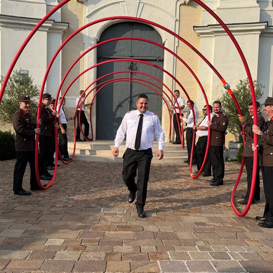 Eine Gruppe von Männern in Uniformen mit roten Rohren vor einem Gebäude. Ein Mann in der Mitte, der ein weißes Hemd und eine schwarze Krawatte trägt, lächelt.