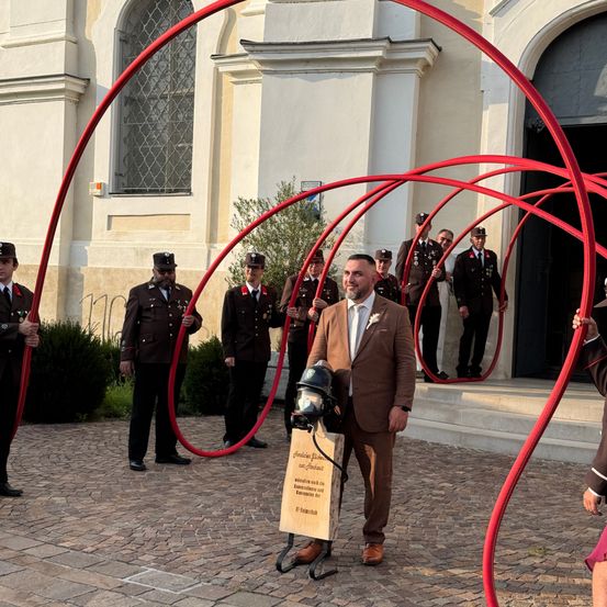 Ein Mann in einem braunen Anzug, der ein Schild hält, steht vor einer Gruppe uniformierter Offiziere. Sie halten rote Gegenstände vor einem Gebäude.