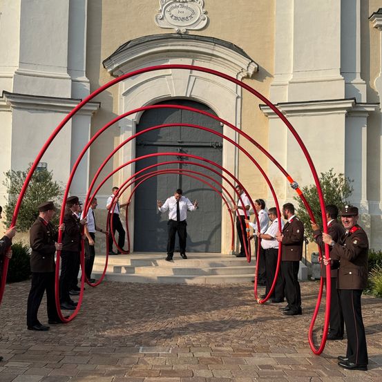 Eine Gruppe uniformierter Personen führt eine Zeremonie im Freien vor einem Gebäude mit einem gewölbten Eingang durch. Sie halten rote Rohre.