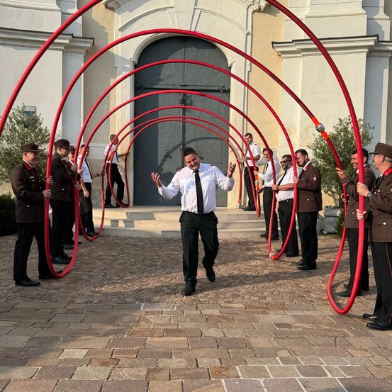 Eine Gruppe uniformierter Personen führt eine koordinierte Aktivität mit roten Rohren vor einem Gebäude durch.