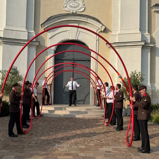 Eine Gruppe uniformierter Personen, die rote Rohre halten, steht vor einem Gebäude mit einem gewölbten Eingang und einem Mann in der Mitte.