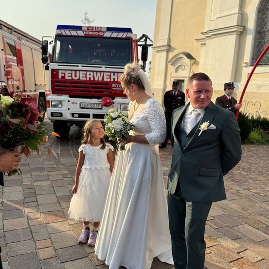 Eine Braut in einem weißen Kleid steht neben einem Bräutigam in einem schwarzen Anzug und hält einen Blumenstrauß. Ein kleines Mädchen in einem weißen Kleid steht zwischen ihnen. Ein Feuerwehrauto ist hinter ihnen geparkt.