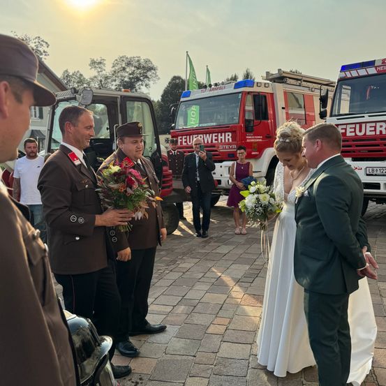 Eine Hochzeitszeremonie findet statt, bei der ein Paar vor Feuerwehrautos steht und zwei Männer Blumen halten.