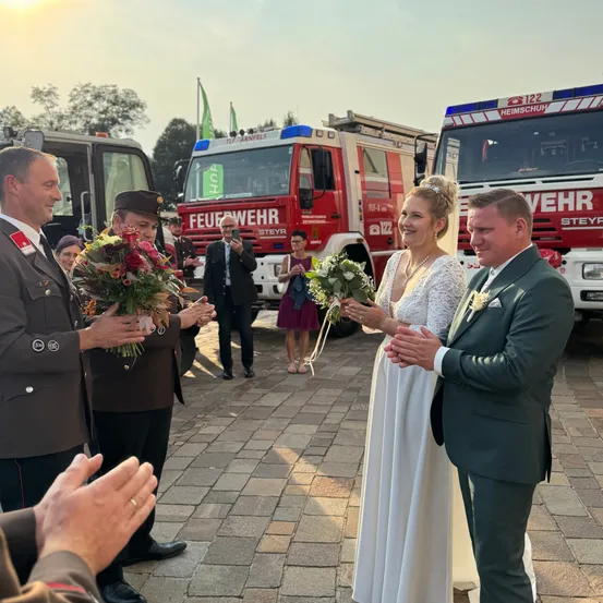Eine Hochzeitszeremonie findet im Freien statt, mit Feuerwehrautos im Hintergrund. Die Braut und der Bräutigam sind von Menschen umgeben, einige halten Blumen. Der Bräutigam trägt einen grünen Anzug, und die Braut hält einen Blumenstrauß. Ein anderer Mann klatscht in der Nähe.