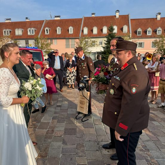 Eine Hochzeitszeremonie findet im Freien statt. Eine Braut in einem weißen Kleid hält einen Blumenstrauß und unterhält sich mit einem Mann in Uniform. Dahinter versammelt sich eine Menge, und ein roter Feuerwehrwagen ist geparkt.