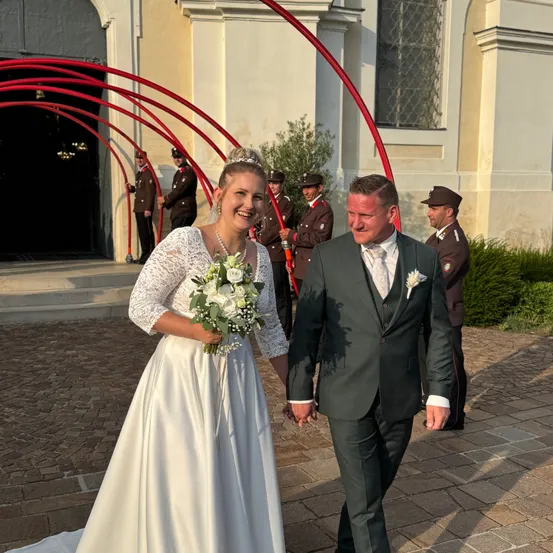Eine Braut und ein Bräutigam gehen Hand in Hand vor einer Kirche, mit uniformierten Wachen im Hintergrund.