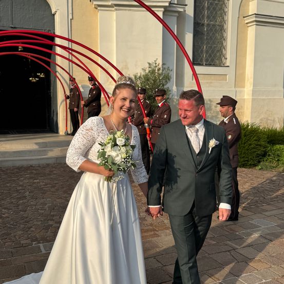 Eine Braut und ein Bräutigam gehen Hand in Hand vor einer Kirche, mit uniformierten Wachen im Hintergrund.