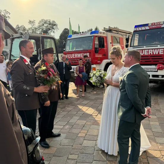Eine Braut und ein Bräutigam stehen vor zwei Feuerwehrfahrzeugen, wobei einer der Männer Blumen hält und der andere sie ansieht. Hinter ihnen versammeln sich mehrere Personen in der Nähe der Feuerwehrfahrzeuge, darunter ein Mann, der ein Foto macht.