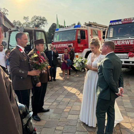Eine Braut und ein Bräutigam stehen vor zwei Feuerwehrfahrzeugen, wobei einer der Männer Blumen hält und der andere sie ansieht. Hinter ihnen versammeln sich mehrere Personen in der Nähe der Feuerwehrfahrzeuge, darunter ein Mann, der ein Foto macht.