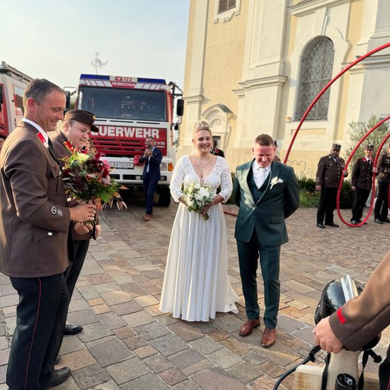 Ein frisch verheiratetes Paar steht vor einer Kirche, ein Feuerwehrmann überreicht einen Blumenstrauß. Ein Feuerwehrauto ist im Hintergrund.